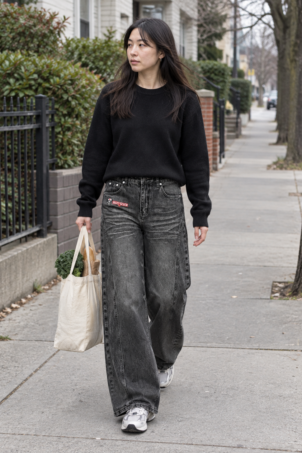 A woman walking on a city sidewalk wearing a black crewneck sweatshirt, wide-leg faded black jeans with a red and white patch on the pocket, and white sneakers, while carrying a cream canvas tote bag.