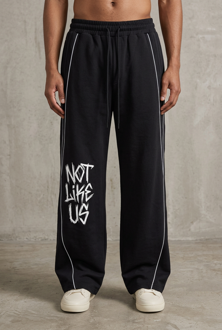 A person wearing black wide-leg sweatpants with white side stripes and a white graphic reading 'NOT LIKE US' on one leg, paired with white sneakers against a textured gray wall.
