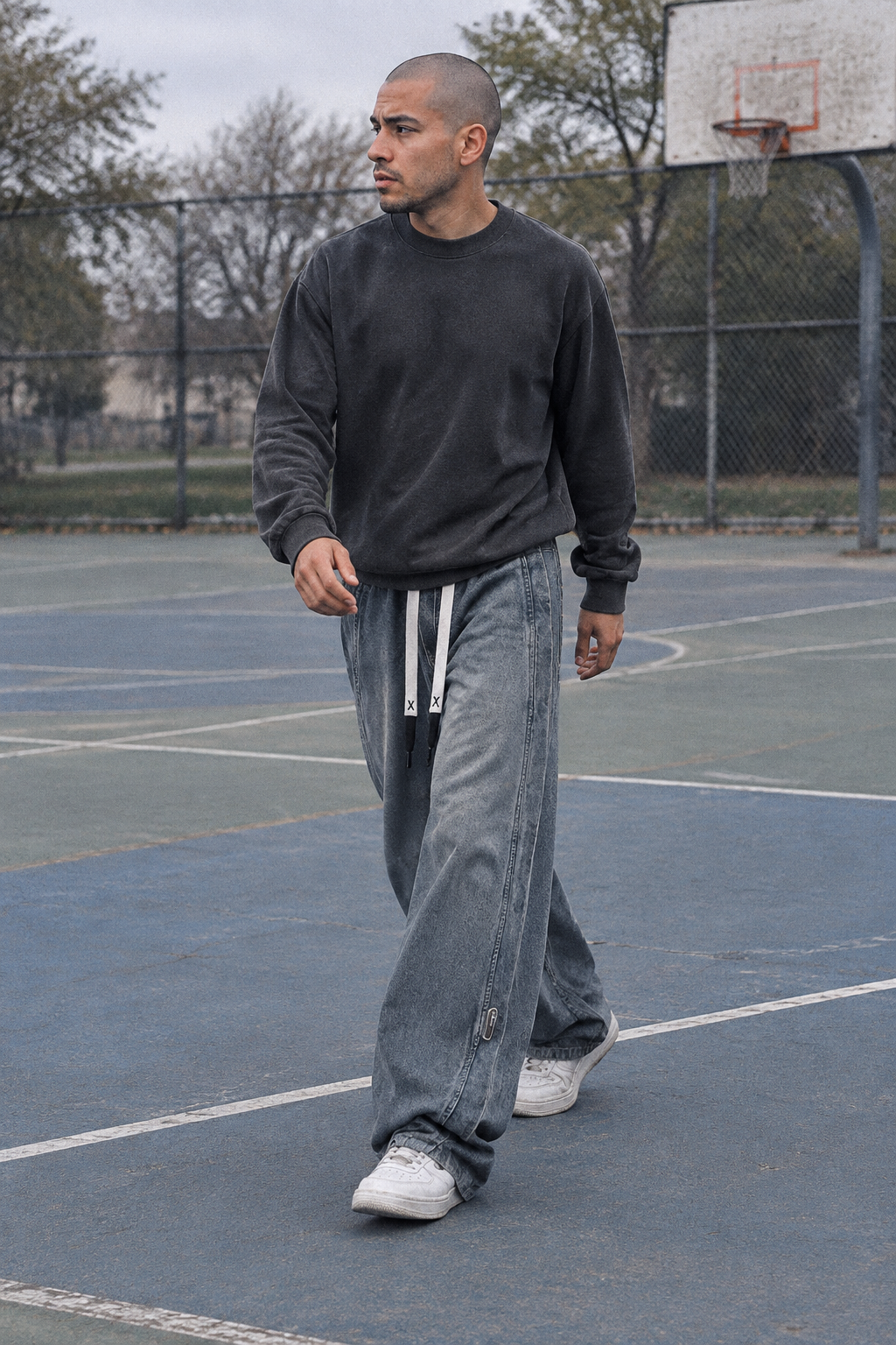 A man walking on an outdoor basketball court wearing a dark charcoal crewneck sweatshirt, wide-leg light-wash jeans with white side stripes, and white sneakers.