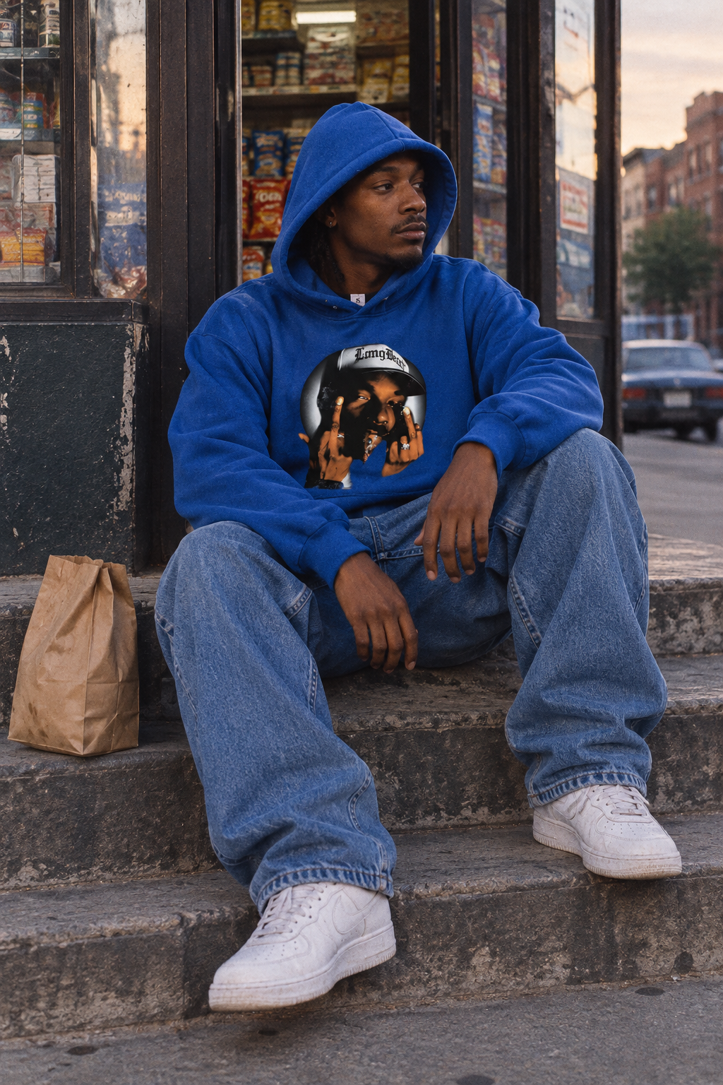 Person wearing the Snoop Dogg graphic hoodie in Color 8 seated outside a corner shop in soft daylight.