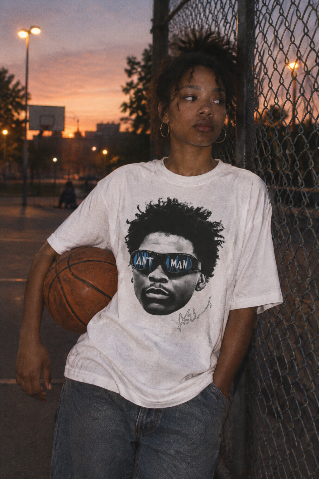 Woman in an oversized white basketball graphic tee leaning on a chain-link fence at dusk holding a basketball