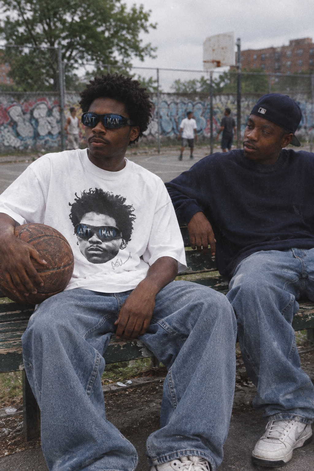 Two friends on a bench by a city basketball court, one wearing an oversized white basketball graphic tee and holding a basketball